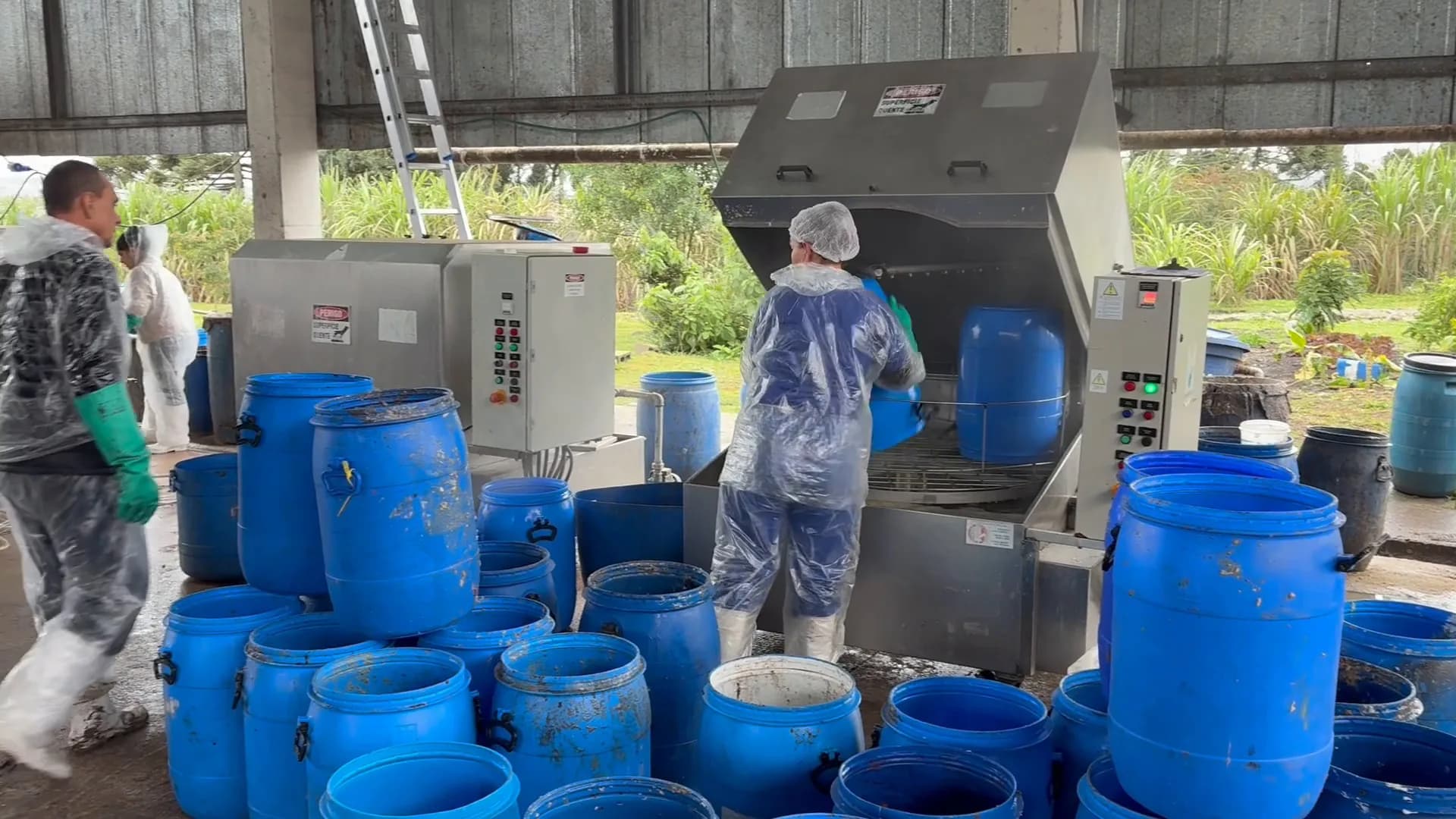 Dois trabalhadores junto à estrutura de lavagem, cercados por bombonas azuis.