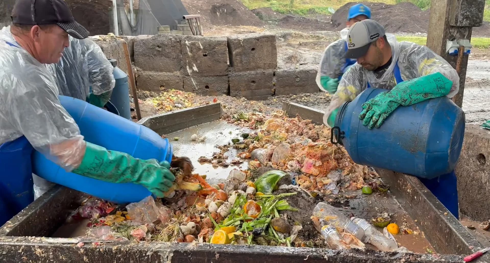 Trabalhadores com luvas verdes separam resíduos de alimentos sobre uma mesa, ao lado de bombonas azuis.