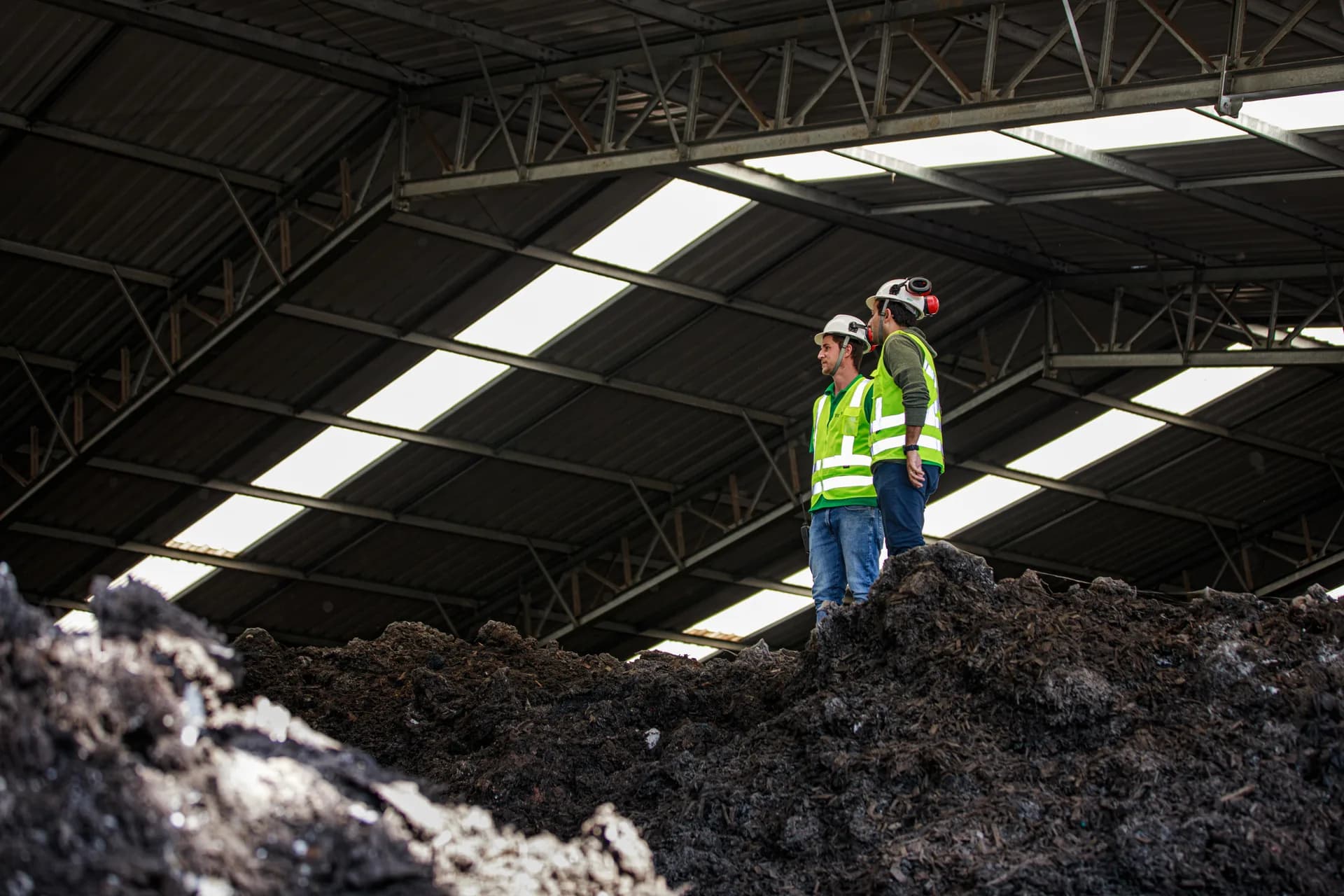 Two workers wearing helmets and reflective vests stand on a compost pile inside the facility.